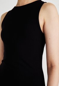 Juodas be rankovių suknelė su lygia, prigludusia forma. Savybės - aukštas kaklas ir minimalūs siūlai, pabrėžiantys elegantišką ir išskirtinai tiesią siluetą.