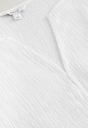 Hașău de țesătură albă cu textură, decolteu în V și etichetă de brand „NEXT” cu inscripția „Fabricat în Turcia”.