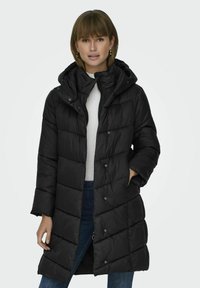 Manteau rembourré noir avec capuche, comportant des sections matelassées, des fermetures à pression à l'avant et des poches latérales. Le tissu semble lisse et isolant.