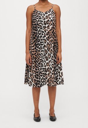 Vrouw draagt een mouwloze jurk tot op de knie met een luipaardprint in bruin, zwart en wit, gecombineerd met zwarte platte schoenen.