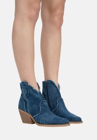 Botas de tobillo de denim con punta afilada, bordes deshilachados, cremallera lateral y tacón apilado. Color: azul oscuro. Presenta costuras decorativas.