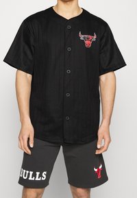 Camiseta negra a rayas con botones en la parte delantera y logo de toro rojo bordado; combinada con pantalones cortos grises con la palabra "BULLS" en blanco.
