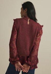 Pull en tricot bordeaux avec un col côtelé et un détail en forme de clé à l'arrière, superposé sur un chemisier floral transparent avec des manches volantées.