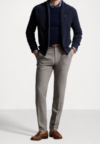 Marineblauwe sweater met ritssluiting over een donker shirt, gecombineerd met lichtgrijze broek en bruine leren schoenen. Kenmerkt zich door een strak, op maat gemaakt ontwerp.