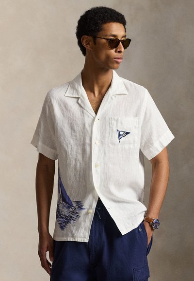 Homme portant une chemise blanche à manches courtes boutonnée avec un imprimé de voiliers, un short bleu marine, des lunettes de soleil et une montre-bracelet, regardant sur le côté.