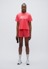 Αθλητικό σετ Nike Pro σε φωτεινό ροζ, με φαρδιά μπλούζα τύπου T-shirt με λευκό λογότυπο και ταιριαστό σορτς. Φοριέται με λευκές κάλτσες και γκρι αθλητικά παπούτσια.