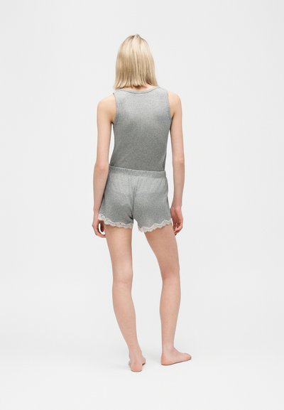 Άτομο με ξανθά μαλλιά που στέκεται ξυπόλυτο, φορά γκρι αμάνικη μπλούζα και ταιριαστά σορτς με λευκή δαντελωτή μπορντούρα, γυρισμένο πλάτη σε απλό φόντο.