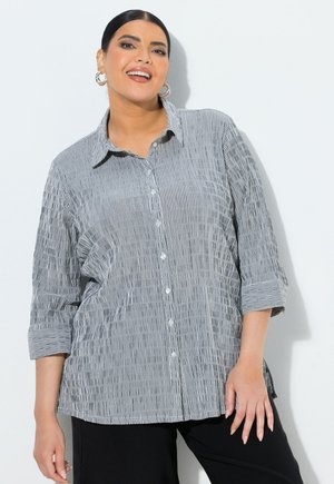 Mujer con cabello oscuro recogido en un moño bajo, lleva pendientes de aro plateados, una camisa de rayas grises y blancas con botones y mangas remangadas, y pantalones negros.