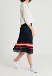 Gonna plissettata blu navy elastica con righe orizzontali rosse e bianche all'orlo, abbinata a un cardigan beige chiaro e sneakers bianche.