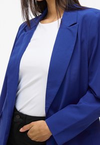 Femme portant un blazer bleu sur une chemise blanche, une main reposant dans la poche d'un pantalon noir, vue du cou à la taille.
