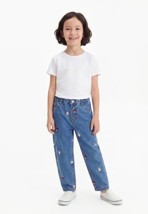 Jeune enfant aux cheveux bruns courts portant un t-shirt blanc, un jean bleu avec broderie florale et des chaussures blanches à enfiler, souriant et debout.