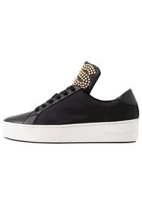 Baskets noires avec dessus en cuir et tissu, présentant un motif à clous dorés sur la languette, une semelle blanche contrastante et un détail de logo sur le côté.