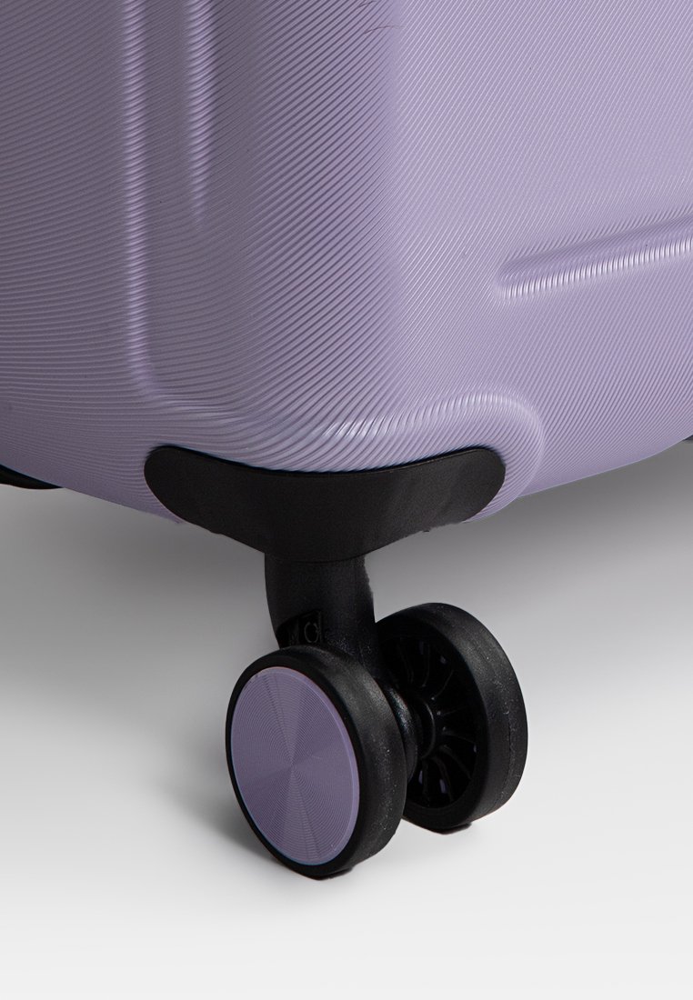 Valise rigide lavande avec des lignes verticales et une base noire. Elle est équipée d'une seule roue noire avec un accent lavande sur le rebord.