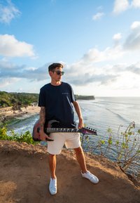 Čierna elektrická gitara držaná osobou v námorníckom tričku a bielych šortkách, stojacou na skalnatom výbežku s plážou a oceánom v pozadí.