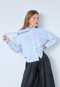 Blusa de algodón con rayas verticales en azul claro y blanco, cuello abotonado, mangas largas y corte holgado, combinada con pantalones oscuros con pliegues.
