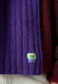 Lila pletena jopica z ribasto teksturo, z oznako na spodnjem levem delu. Ozadje ima bordo flisasto tkanino.