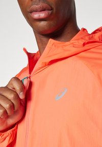 Veste légère orange avec fermeture éclair à l'avant, dotée d'une capuche et d'un logo réfléchissant sur la poitrine. Texture en nylon lisse avec poignets élastiques.