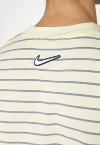 Ljust gul kortärmad skjorta med horisontella grå ränder och en liten marinblå Nike-logotyp broderad på baksidan nära kragen.
