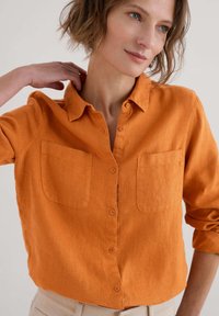 Oranžová lněná košile s knoflíkovým zapínáním vpředu, dvěma náprsními kapsami a ohrnutými rukávy. Textura vypadá měkce a prodyšně.