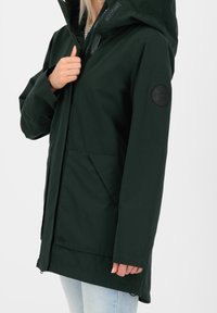 Tmavě zelená, dlouhá bunda s kapucí, se zipovým zapínáním, elastickými manžetami a kulatou černou nášivkou na horní části rukávu. Obsahuje přední kapsy.