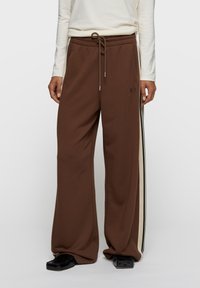 Pantalon de survêtement marron à jambes larges avec taille à cordon, présentant des bandes latérales crème et noires ainsi qu'un petit logo à l'avant.