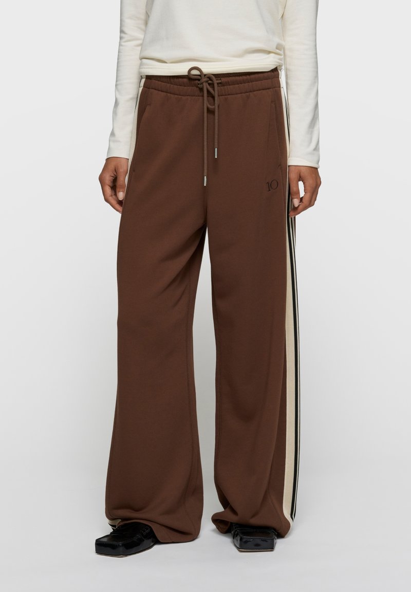 Pantalon de survêtement marron à jambes larges avec taille à cordon, présentant des bandes latérales crème et noires ainsi qu'un petit logo à l'avant.