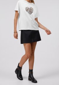 Woman wearing white T-shirt with leopard heart print, black leather mini skirt, black studded ankle boots, and gold bracelet, standing on gray background.