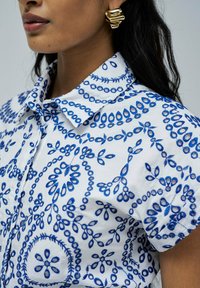 Camisa blanca con patrones florales bordados en azul, cuello con solapa y mangas cortas. Pendientes abstractos dorados añaden detalle.