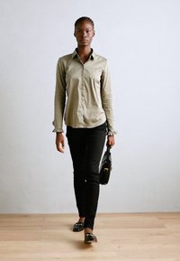 Camisa de mangas longas verde-oliva com fecho de botão, jeans skinny pretos e bolsa preta, em pé sobre um chão de madeira clara contra uma parede cinza claro.