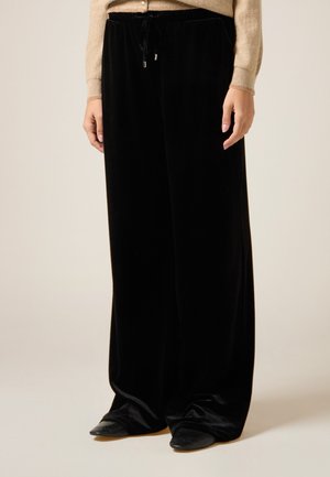 Černé sametové kalhoty s širokými nohavicemi a elastickým pasem, s šňůrkou na stažení a měkkou texturou, doplněné o špičaté černé boty.