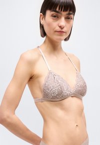 Νεαρή γυναίκα με κοντά σκούρα μαλλιά που φορά σκουρό ροζ δαντελένιο σουτιέν και ταίριαστο εσώρουχο, στέκεται μπροστά σε απλό φωτεινό φόντο.