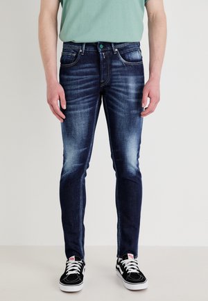 Tamsiai mėlynos džinsinės kelnės su siauru siluetu, su išblukusiu rankiniu plovimu, standartinėmis kišenėmis ir mygtuku užsegimu. Derinamos su juodais sportiniais bateliais.