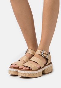 Plattform sandaler med vävda beige remmar, en tjock sula med texturerad yta och metallspännen för justering.