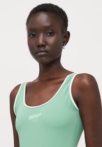 Maillot de bain vert menthe avec des bordures blanches, arborant le logo "Hilfiger" au centre. Texture lisse et design athlétique pour une coupe ajustée.