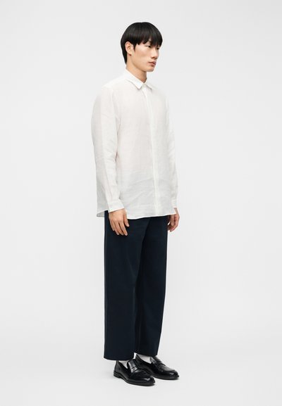 Chemise blanche en lin à boutons, coupe ample, manches longues, portée avec un pantalon large bleu marine foncé et des chaussures en cuir noir.