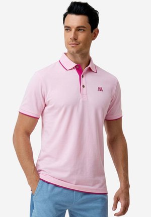 Mann trägt ein hellrosa Poloshirt mit dunkelrosa Besatz und blaue Hose, steht mit einer Hand in der Tasche und schaut leicht nach rechts.