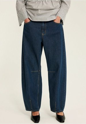 Person iført mørkeblå vidde jeans med frontsøm detaljer, hænderne i lommerne, stylet med sorte sko med firkantet tå og en stribet skjorte.