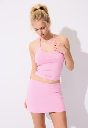 Młoda kobieta z blond włosami związanymi w kucyk, ubrana w jasnoróżowy top halter i dopasowaną krótką spódniczkę na jednolitym białym tle.