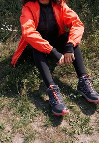 Zwarte wandelschoenen met rode accenten, gecombineerd met zwarte leggings en een oranje windjack, zittend op gras.