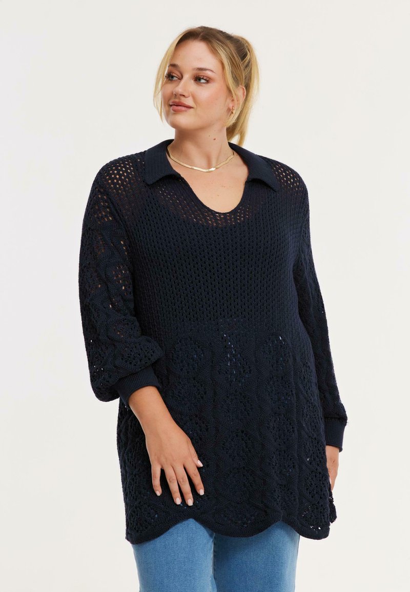 Frau mit blondem Haar, die einen lockeren marineblauen Strickpullover mit Kragen und blaue Jeans trägt und zur Seite schaut.