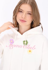 Fehér kapucnis pulóver, "Mymo Club" felirattal rózsaszín betűkkel, és három texturált virágapplikációval lila, sárga és zöld színekben. Húzózsinóros kapucni.