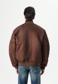 Casaco puffer castanho com gola alta, punhos e bainha canelados; apresenta costura de logo discreta nas costas, combinado com jeans de ganga azul.