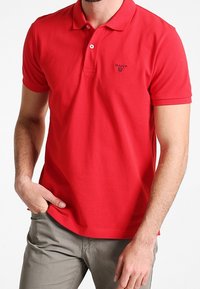 Rød bomulds polo-shirt med krave, tre knapper og et lille sort logo på brystet. Båret med lyse bukser.