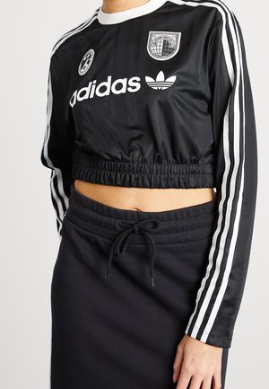 Czarny krótki top Adidas z długim rękawem, z białymi paskami na rękawach, logo i naszywkami, w połączeniu z czarną spódnicą z troczkiem.