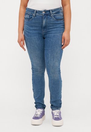 Jeans skinny em denim azul com uma cintura alta, cinco bolsos e uma textura suave. Usados com ténis roxos com cadarços brancos.