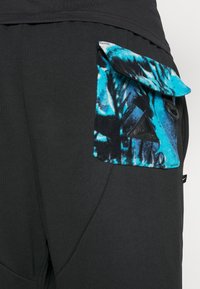 Schwarze Jogginghosen mit einer strukturierten, bunten Tasche, die blaue und schwarze Muster aufweist. Enthält ein dreieckiges Logodetail in der Nähe der Tasche.