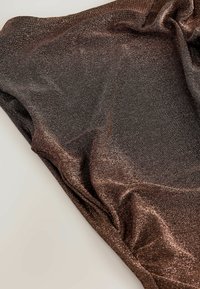 Třpytivá hnědá látka, hladká textura, s jemným leskem. Materiál je zavěšený, ukazuje sklady a různorodé odrazy na povrchu.