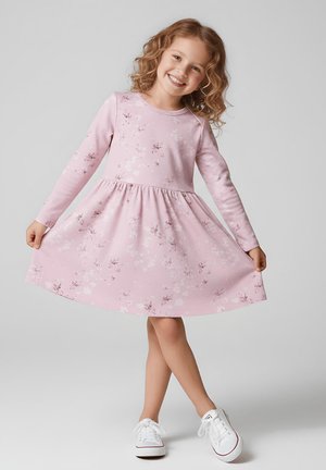 Uśmiechnięta dziewczyna w różowej kwiecistej sukience z długim rękawem i białych trampkach, trzymająca rąbek sukienki i stojąca z przekroczonymi nogami na szarym tle.