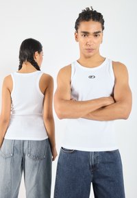 White ribbed tank top with a round neckline and a small black logo at the center. Shown on two models wearing loose denim pants.