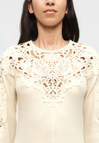 Γυναίκα που φοράει κρεμ πλεκτό τοπ με περίτεχνη δαντέλα στον λαιμό και τα μανίκια, δείχνοντας το ανώτερο μέρος του κορμού και το λαιμό.